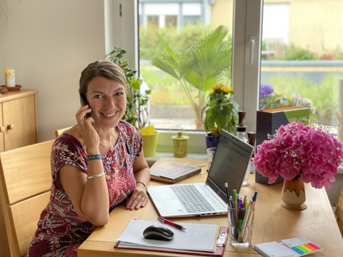 Frau sitzt am Küchentisch und telefoniert, vor ihr steht ein Laptop.