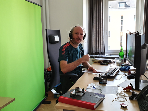 Professor sitzt vor Greenscreen am Schreibtisch.