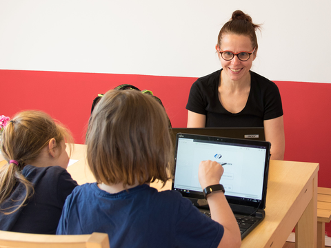 Frau sitzt zwei Kindern am Laptop gegenüber und macht mit ihnen Hausaufgaben.