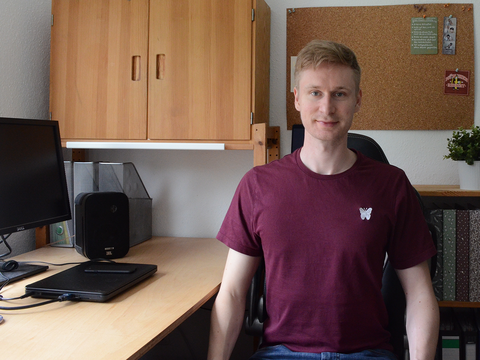 Student schaut in Kamera und sitzt zu Hause vor seinem Schreibtisch mit Computer.