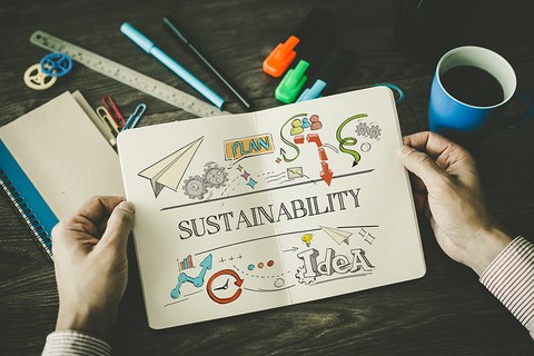 Ein Arbeitsplatz, über dem zwei Hände ein Notizbuch halten mit einer Skizze zum Thema "Sustainabilty".