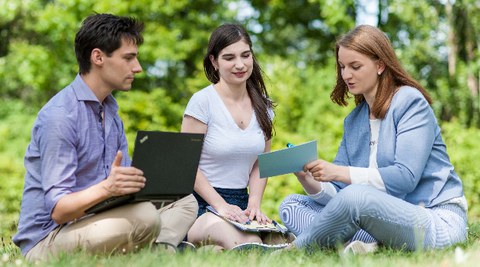 Zu sehen ist ein Foto von drei Studierenden, welche auf einer Wiese sitzen. Links im Bild sitzt ein junger Mann mit einem Laptop auf dem Schoß. In der Mitte und rechts sitzen zwei junge Frauen. Die Frau rechts hält eine Karteikarte in der Hand.