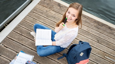 Eine junge Frau lacht in die Kamera. Sie sitzt auf einem Steg am Wasser und hält einen Stift und ein Buch in ihren Händen.