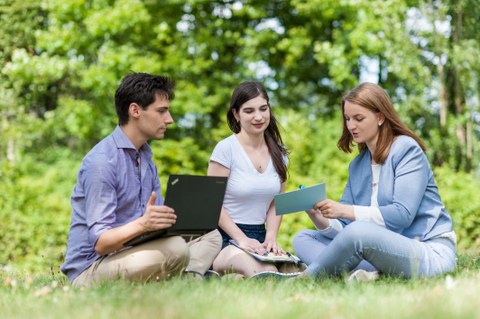 Zu sehen ist ein Foto von drei Studierenden, welche auf einer Wiese sitzen. Links im Bild sitzt ein junger Mann mit einem Laptop auf dem Schoß. In der Mitte und rechts sitzen zwei junge Frauen. Die Frau rechts hält eine Karteikarte in der Hand.