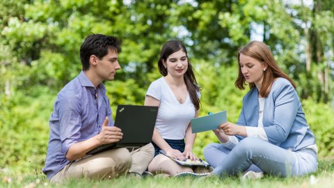 Zu sehen ist ein Foto von drei Studierenden, welche auf einer Wiese sitzen. Links im Bild sitzt ein junger Mann mit einem Laptop auf dem Schoß. In der Mitte und rechts sitzen zwei junge Frauen. Die Frau rechts hält eine Karteikarte in der Hand.