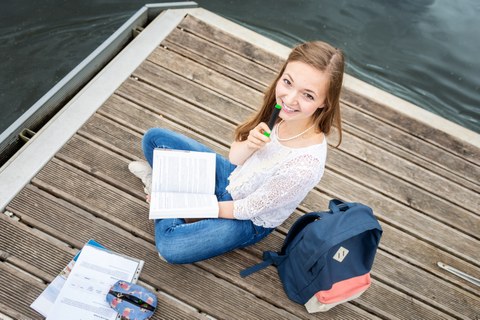 A young woman laughs into the camera. She is sitting on a jetty by the water and holds a pen and a book in her hands.
