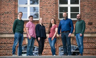 2 women and 4 men pose for a group picture infront af a brick building with white windows