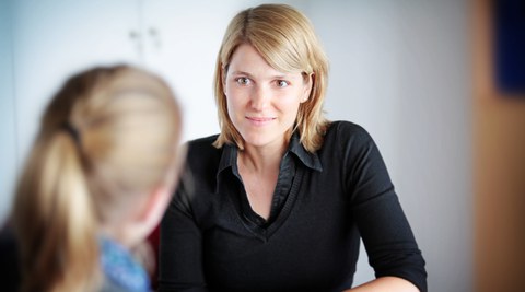 Zu sehen ist ein Foto von zwei Personen an einem Tisch. Im Vordergrund ist leicht verschwommen eine Person von hinten zu erkennen. Ihr gegenüber sitzt eine Studienberaterin.