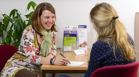 Es ist ein Foto zu sehen, auf dem zwei Personen abgebildet sind. Sie sitzen sich gegenüber an einem Tisch. Die Person links im Bild ist eine Studienberaterin, welche sich Notizen macht. Die zweite Person ist nur von hinten zu erkennen.