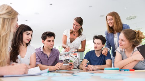 Auf diesem Foto sind mehrere Personen während eines Workshops zu sehen. Fünf Studierende sitzen an einem großen Tisch, auf welchem unterschiedliche Büroutensilien liegen. Zwei Frauen stehen am Tisch und wenden sich den Studierenden zu. 