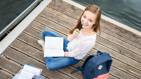 A young woman laughs into the camera. She is sitting on a jetty by the water and holds a pen and a book in her hands.