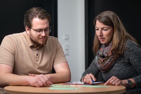 Eine Frau und ein Mann sitzen zueinander gewandt an einem runden Tisch und unterhalten sich. Es handelt sich um eine Studienberaterin und einen Klienten.