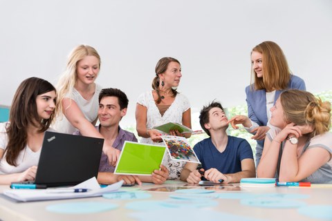 This photo shows several people during a workshop. Five students are sitting at a large table on which various office utensils are lying. Two women stand at the table and turn to the students.