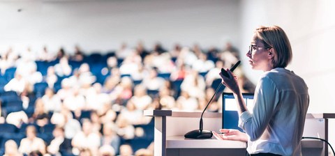 Foto: Eine Person spricht an einem Rednerpult vor einem Publikum in einem Konferenzraum