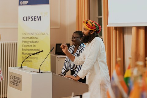 Ein Mann mit Tuch um den Kopf gebunden und eine Frau stehen gemeinsam an einem Rednerpult. Im Hintergrund ein Aufsteller mit der Aufschrift "Cipsem"