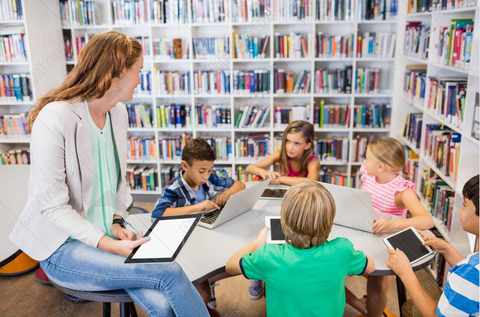 Foto einer Lehrerin, die mit einem tablet in der Hand auf einem Tisch sitzt. An dem Tisch lernen 5 Grundschulkinder.