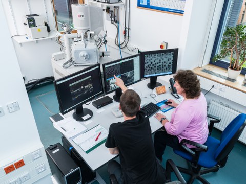 zwei Personen sitzen mit an einem Schreibtisch mit drei Monitoren. Im Hintergrund stehen größere technische Geräte.