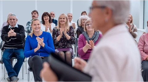 Foto von älteren Menschen in einer Seminarveranstaltung, im Vordergrund eine ältere Dame, die anderen Menschen klatschen