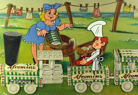 The photo shows two animated figures. A woman washes laundry on a washboard. A man with a chef's hat and apron stands next to her. In the foreground: a train made of breath mint boxes.