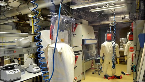 The photo shows a laboratory with various machines and a number of hazmat suits.