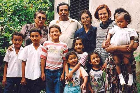 The photo shows Anja Centeno García surrounded by her host family. We see four women, one man, and seven children. They are standing in front of a building and a large bush.