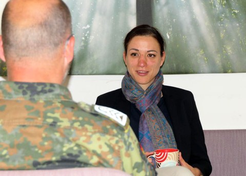 The photo shows Ariane Knechtel facing the camera. In the foreground, we recognize the upper body and the back of a man's head. He is wearing an armed forces uniform and is engaged in conversation with Ariane Knechtel.