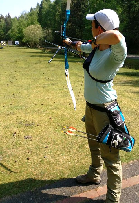 The photo shows Ariane Knechtel practicing archery. She is faced away from the camera and is aiming at a target in the distance.