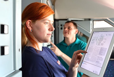 The photo shows a woman studying a blueprint on a tablet. A man is standing in the background.