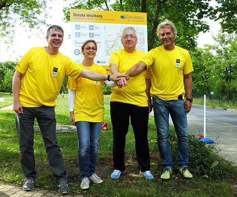 The photo shows four people in yellow t-shirts with their arms outstretched and their hands touching. They are standing outside in front of a sign with a map of a walking trail.