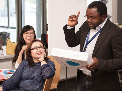Das Foto zeigt Dr. Sefirin Djiogue, der gerade etwas erklärt. Dabei hält er den Zeigefinger nach oben gestreckt. Im Hintergrund sitzen zwei Frauen an einem Tisch.