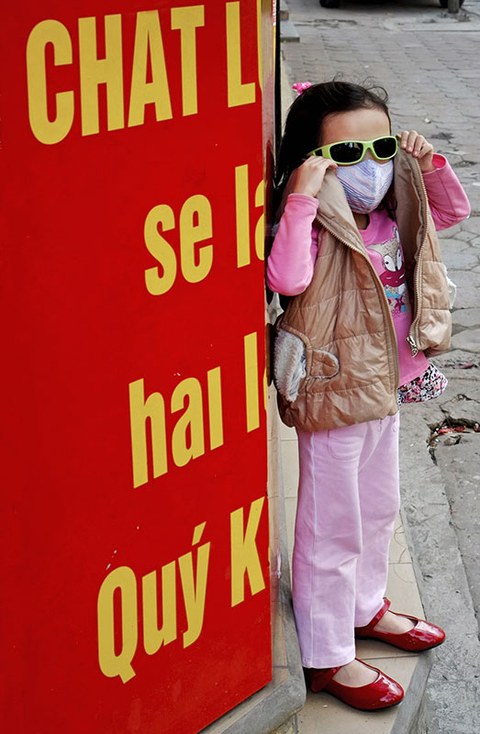 Das Foto zeigt ein kleines Mädchen mit Mundschutz vor einer roten Plakatwand.