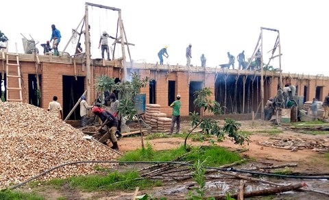 The photo shows the construction of a school in Burkina Faso. Many people work atop the roof while others look on from the ground below.