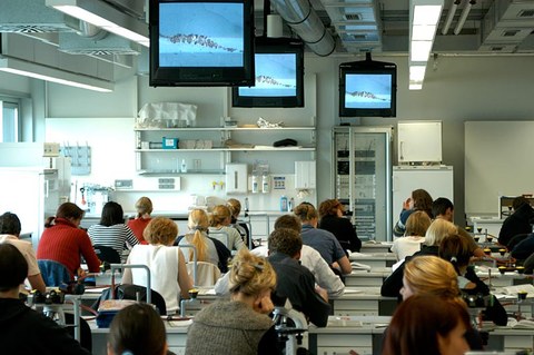 The photo shows students in a classroom at the Faculty of Medicine. They are seated facing the front. We see their backs.