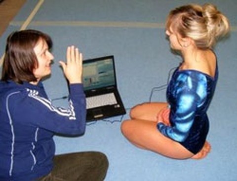 Dr. Reimann explains something to a gymnast. They are seated on a padded floor and there is a laptop in front of them, with wires leading to the gymnast.