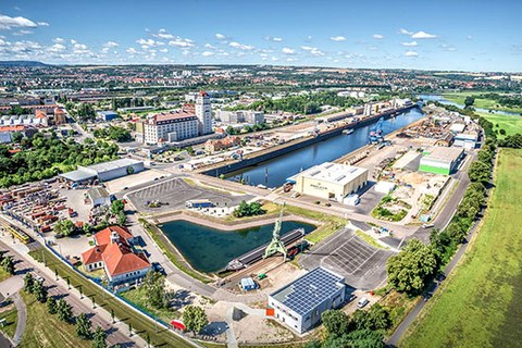 The photo shows the Alberthafen port in Dresden. On the right side of the water we see an industrial setting and then a green field. On the right side there is a cityscape with trees and buildings.