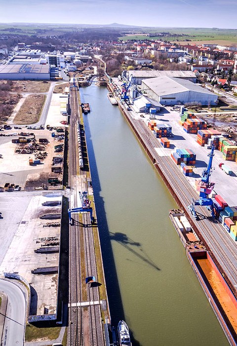 The photo shows an aerial view of the inland port of Riesa. There are tracks running along both sides of the water. On the right side we also see lots of stacked sea containers, a crane and a warehouse.