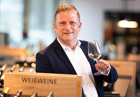 The photo shows Ivo Reuter. He is holding a glass of white wine in his hand and leaning against a shelf labeled "white wines."