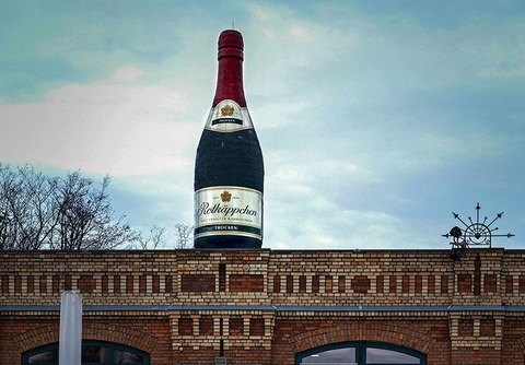The photo shows a large model of a Rotkäppchen sparkling wine bottle atop the roof of a building. The bottle is green and the foil around the top as well as the cork are red.
