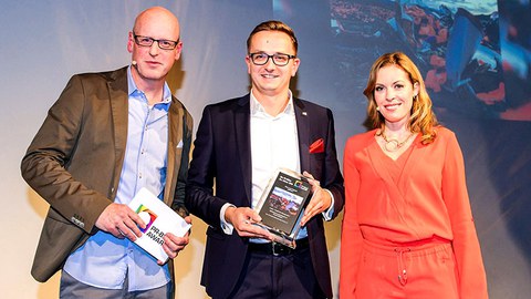 The photo shows three people at an award ceremony. Kai Kranich stands in the center showing the prize to the camera.