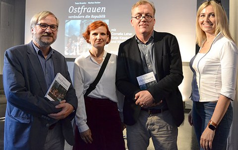 The photo shows four people. They are standing next to each other and smiling at the camera. In the background, there is a presentation titled "Ostfrauen" ("East German Women").