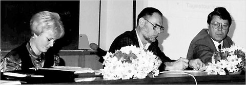 The photo is in black-and-white, and it shows three people sitting at a long table with flowers and table-top microphones.