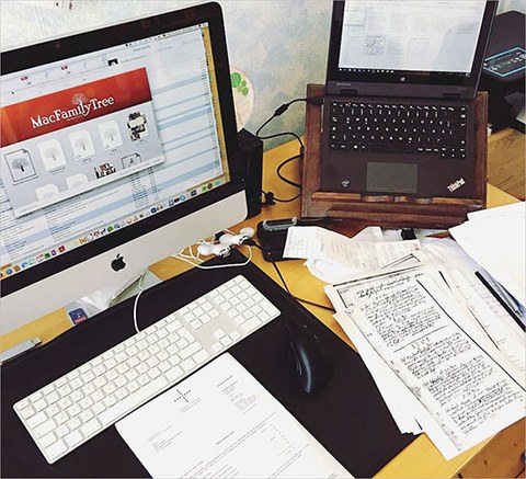 The desk of a professional genealogist. We see a spread of papers, a desktop computer with a family tree and a laptop.