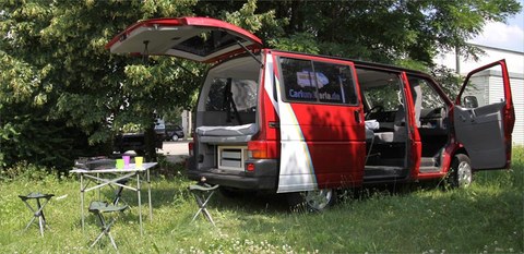 The photo shows a green camping van. A table with chairs stands in front of the van.