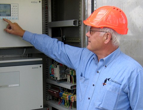The photo shows Ralf Kelm standing in front of an open control cabinet and pointing to the display screen.