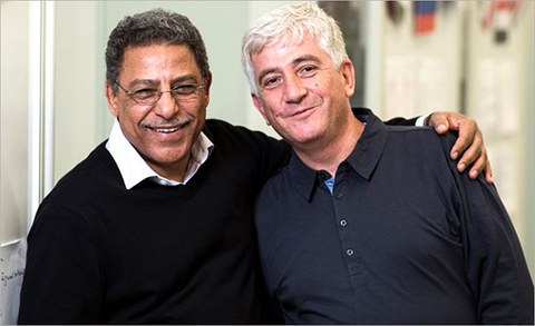 The photo shows two men who are looking at the camera and smiling. The man on the left has his arm around the shoulders of the man on the right.