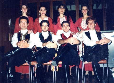 The photo shows a dance ensemble in uniform. Four men sit in chairs before four women.