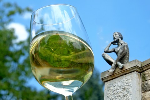 The photo shows a glass of white wine from below in the foreground and a statue of a person sitting on a ledge with their legs crossed and hands under their chin higher up in the background.