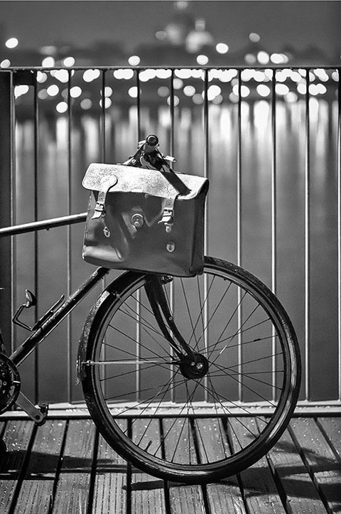 This black-and-white photo shows the front wheel of a bicycle on a bridge with a briefcase hanging from the handlebars. In the background, a river and brightly lit buildings.