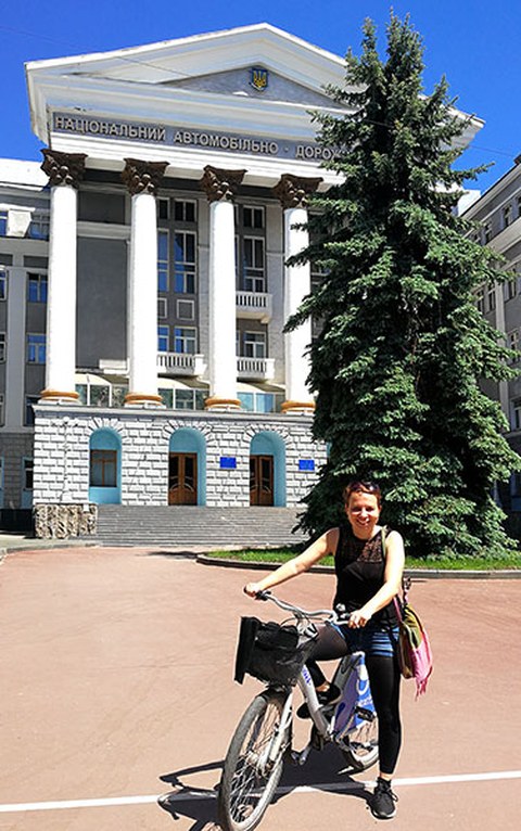 Angela Francke on her bike, standing before a building with large columns. In front of it is a tree. It is a very sunny day with blue skies.