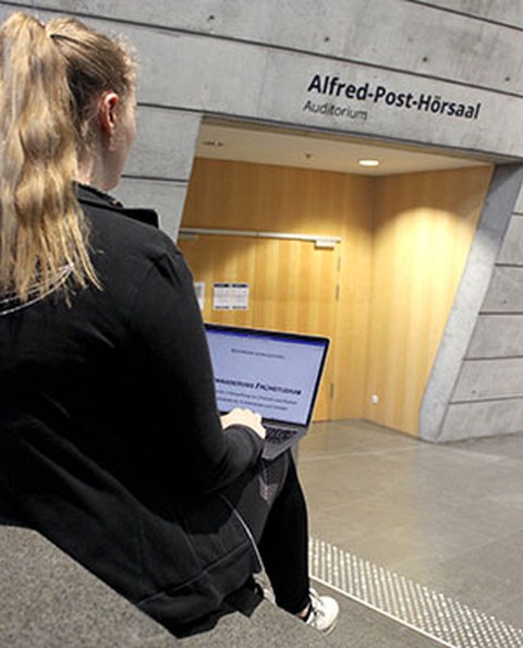 Photo of Stefanie Gentzsch from behind. She is sitting on stairs in front of the entrance to a lecture hall with a laptop on her lap.